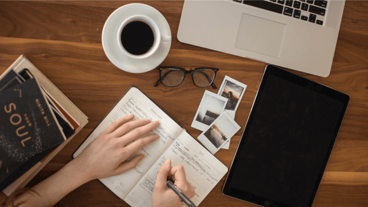 Image of hands writing in an open productivity journal on a wooden desk with additional items including a laptop, cup of coffee, pair of glasses, a closed notebook, a stack of books, and three photos.