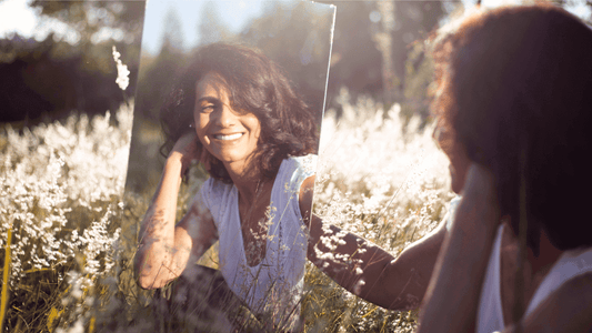 Image of a woman in a field looking at her own reflection in a mirror.
