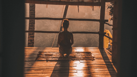 Image of a woman sitting on a balcony at sunrise in Meditation Pose.