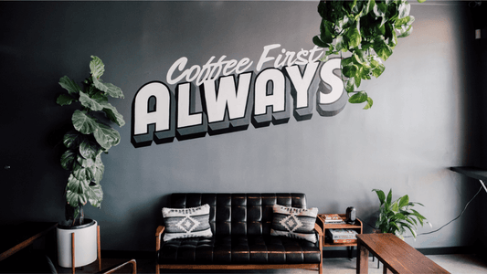Image of a black leather couch with two pillows against a gray wall with writing that says "Coffee First Always". A perfect environment for productivity.
