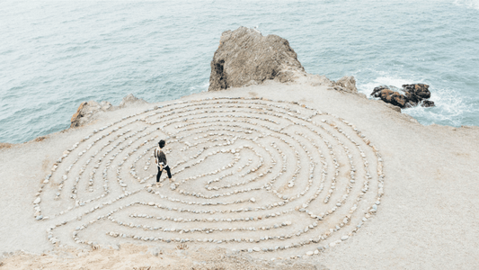 Image of a person walking a labyrinth made of stones in the sand with the ocean in the background, a perfect environment to cultivate self-awareness and mindful planning.