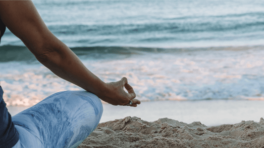 Image of the right side of a person sitting on sand while looking at ocean waves crashing on shore sitting in the meditation pose contemplating habits for a balanced life.