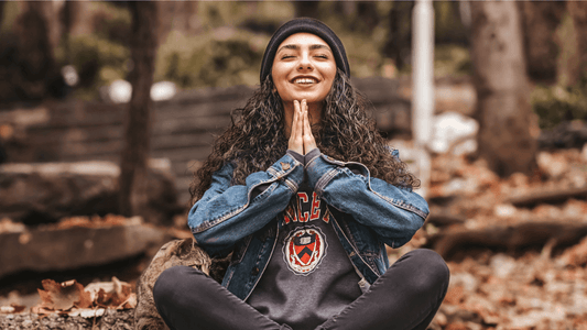 Image of a woman with with long, dark, curly hair wearing black jeans, t-shirt, and jean jacket, smiling with eyes closed while sitting in the woods during fall in the yoga Prayer Pose, taking time thanks to the art of mindful scheduling.